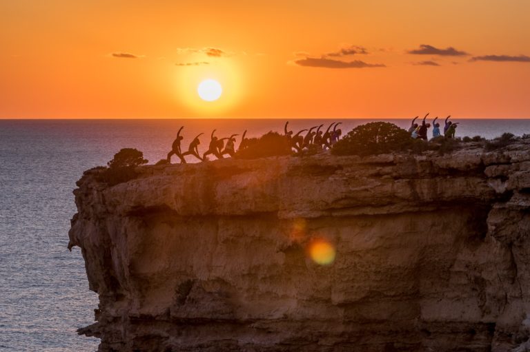 Eine Gruppe von Menschen springt bei Sonnenuntergang silhouettiert auf einer Felsklippe über dem Meer – © Axel Hebenstreit