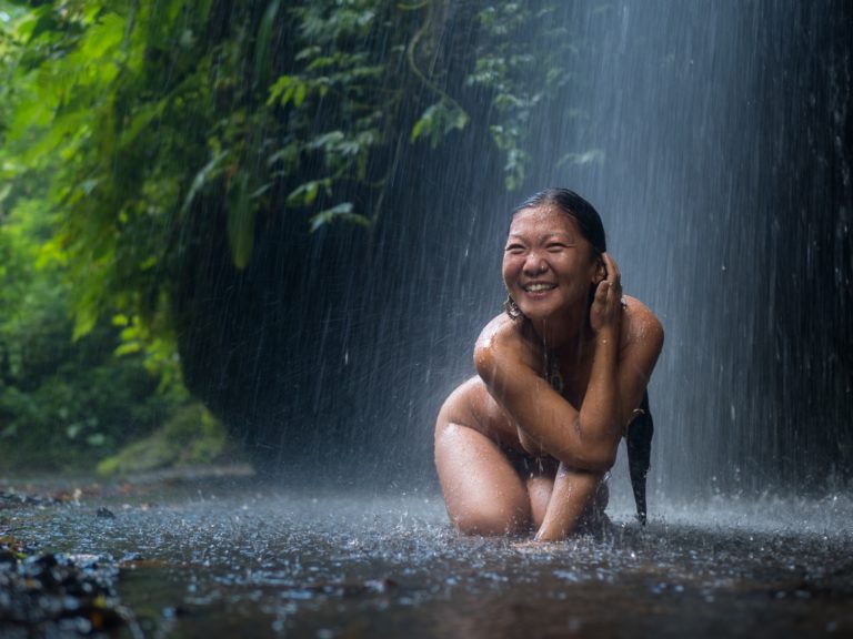 Lächelnde Frau kniet im Regen unter einem Wasserfall im grünen Wald und genießt die erfrischende Naturerfahrung – © Axel Hebenstreit