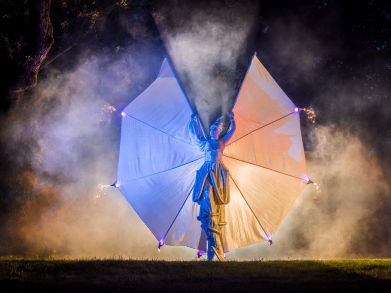 Performer in blauem Kostüm mit leuchtenden Flügeln in Blau und Orange schwebt nachts in Nebelwolken über einer Wiese – © Axel Hebenstreit