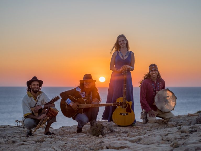Vier Musikerinnen und Musiker mit Gitarren und Rahmentrommel performen bei Sonnenuntergang am Strand mit glühender Sonne im Hintergrund – © Axel Hebenstreit