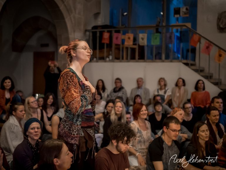 Eine Gruppe von Menschen sitzt zusammen, während eine Frau in bunter Kleidung im Vordergrund spricht und die Aufmerksamkeit auf sich zieht. – © Axel Hebenstreit
