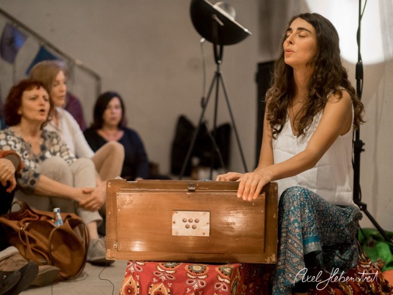 Eine singende Frau sitzt mit einem Harmonium umgeben von Zuhörern in entspannter Atmosphäre. – © Axel Hebenstreit