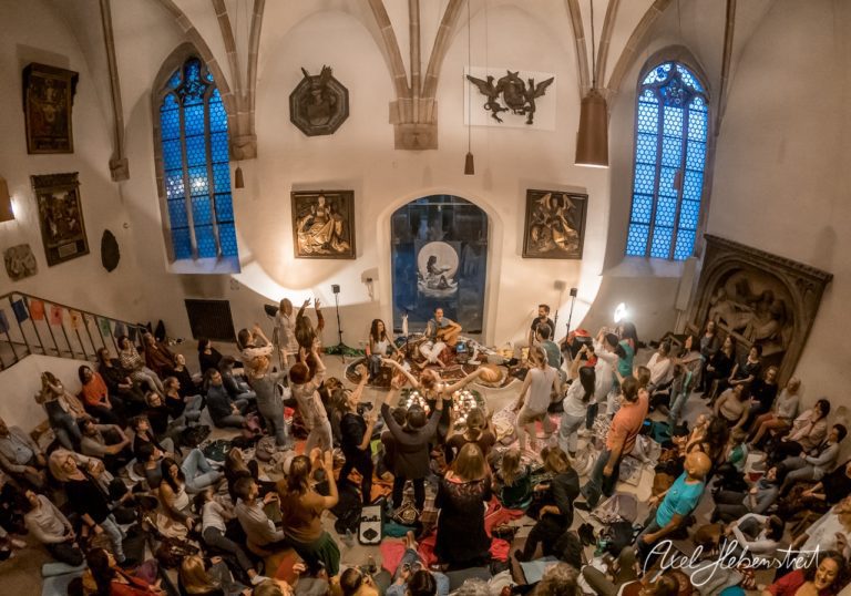 Ein fröhliches Publikum tanzt und musiziert in einem atmosphärischen Raum mit gotischer Architektur. – © Axel Hebenstreit