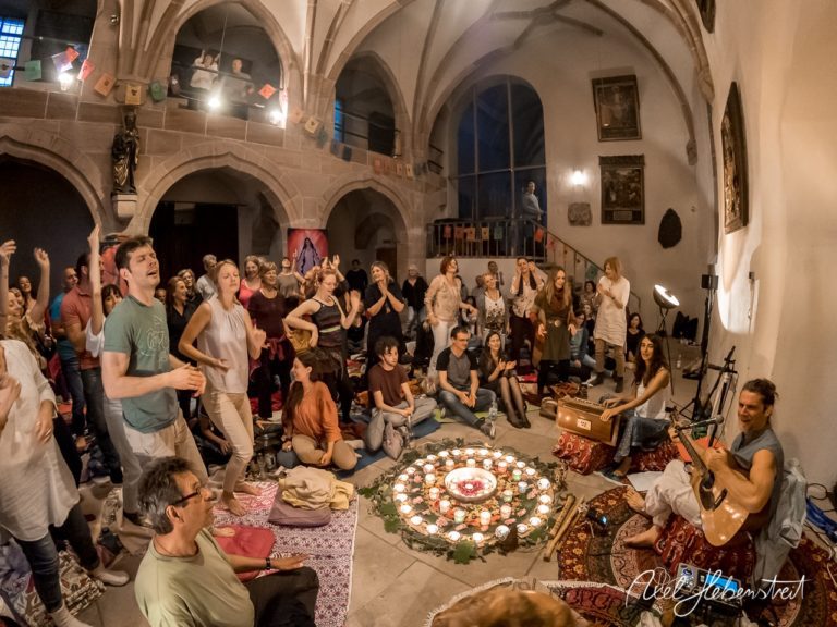 Eine lebendige Gruppe von Menschen tanzt und singt um ein Kerzenlicht-Mandalas im stimmungsvollen Raum. – © Axel Hebenstreit