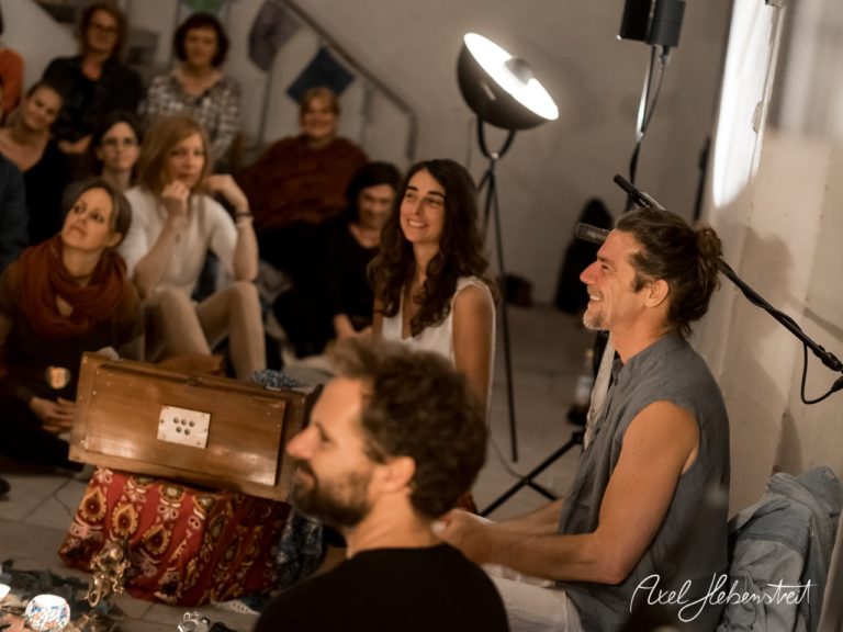 Eine entspannte Gruppe von Menschen lauscht einem Musiker bei einer darbietenden Yoga-Session in einem stilvollen Raum. – © Axel Hebenstreit
