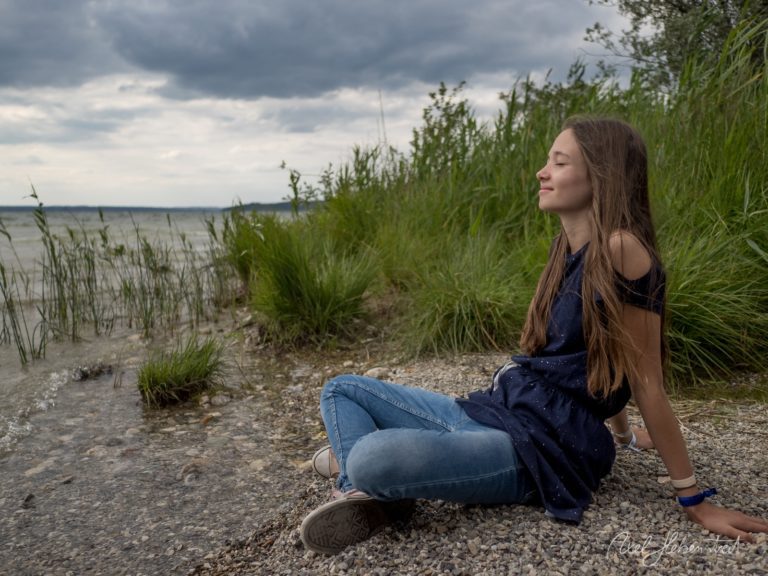 Junge Frau sitzt nachdenklich am Seeufer zwischen Schilf und blickt übers Wasser, bewölkter Himmel – © Axel Hebenstreit