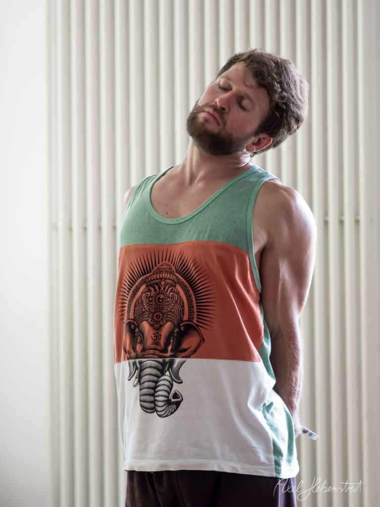 Mann mit Bart in farbigem Tank-Top mit Elefanten-Motiv blickt nachdenklich nach oben vor heller Jalousie – © Axel Hebenstreit