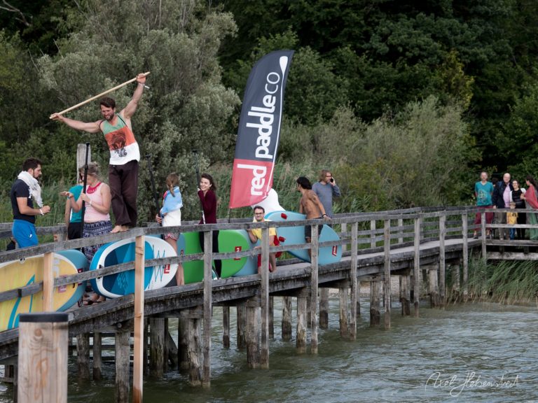 Paddler demonstriert Stand-up-Paddling-Technik auf Steg mit Zuschauern und Bäumen im Hintergrund – © Axel Hebenstreit