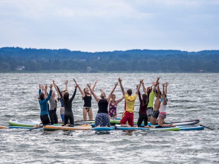 Gruppe jubelnder SUP-Sportler auf ihren Boards im See mit bewaldeten Bergen im Hintergrund – © Axel Hebenstreit