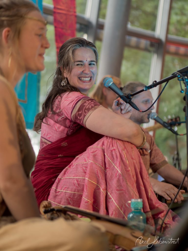 Lachende Sängerin in rosa Gewand sitzt am Mikrofon während eines Festivals in einer Halle mit grünem Ausblick – © Axel Hebenstreit
