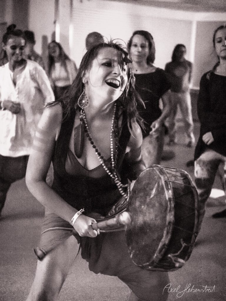 Frau mit Perlenkette spielt Trommel beim Fest, umgeben von feiernden Gästen in Innenraum – © Axel Hebenstreit