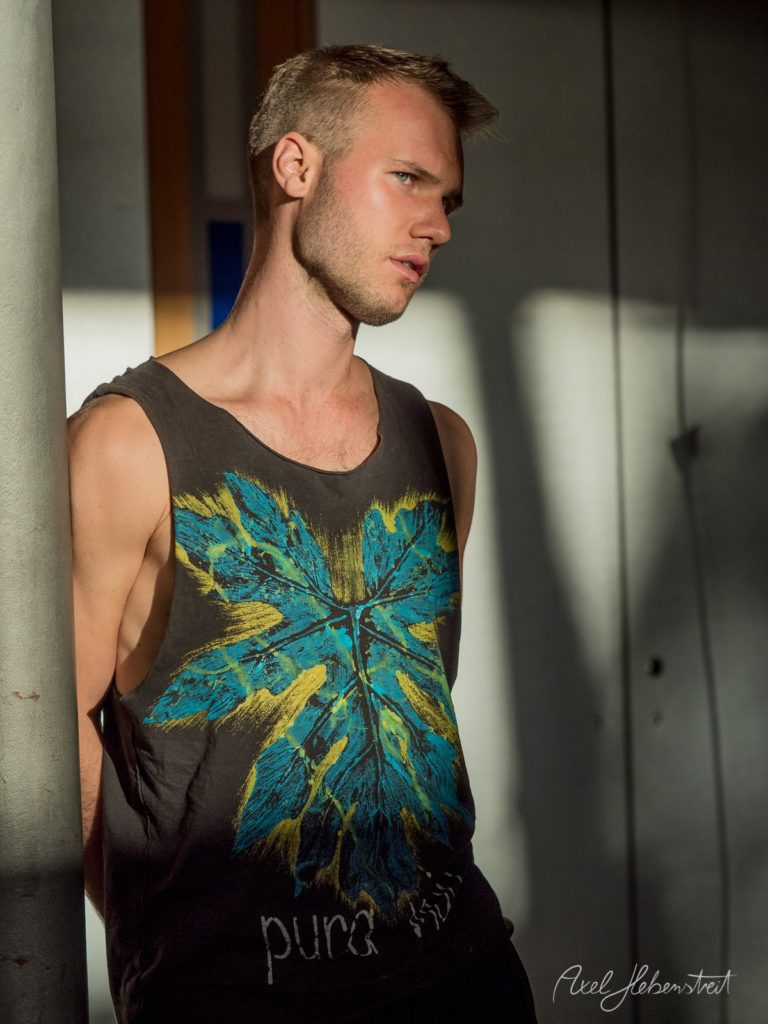 Mann in schwarzem Tanktop mit blauem und gelbem Schmetterlingsmotiv lehnt an grauer Wand in modernem Innenraum – © Axel Hebenstreit