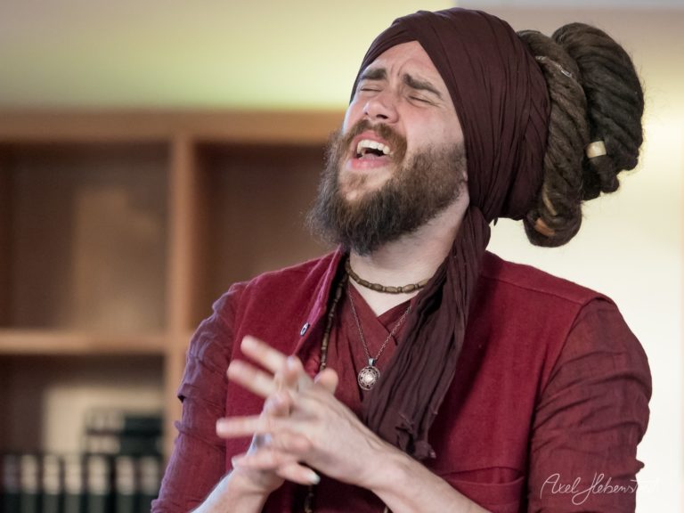 Bärtiger Mann mit dunkelrotem Kopftuch singt leidenschaftlich auf einer Bühne – © Axel Hebenstreit