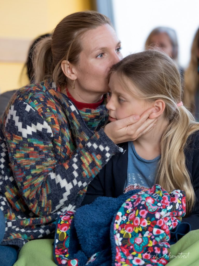 Frau küsst lächelndes Mädchen zärtlich auf die Wange in warmer Innenraum-Atmosphäre – © Axel Hebenstreit