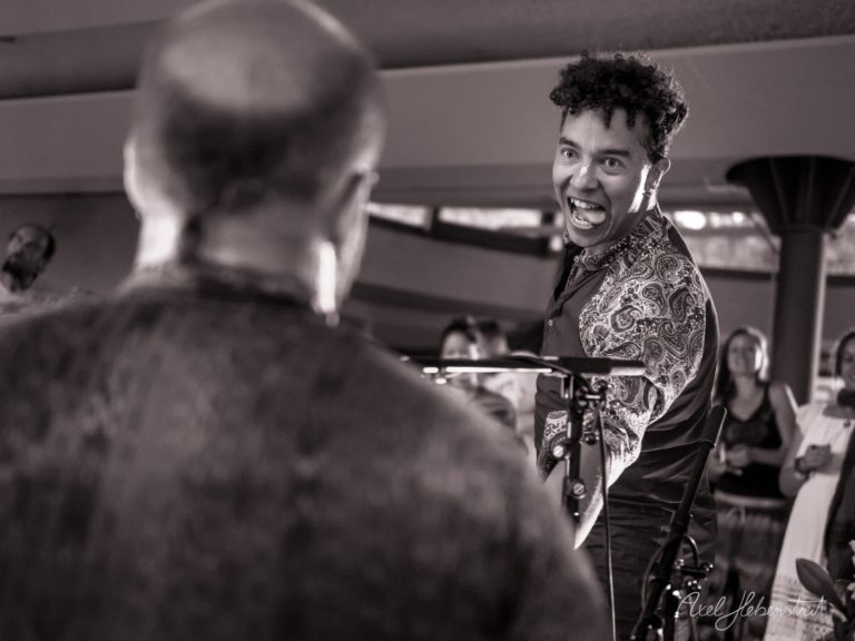 Lächelnder Musiker mit Paisley-Jacke am Mikrofon während eines Indoor-Konzerts mit Publikum – © Axel Hebenstreit