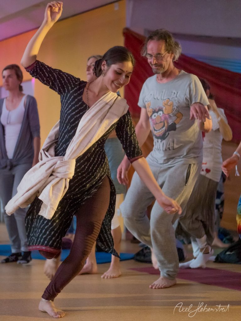 Frau tanzt ausgelassen in schwarzem Outfit während Gruppe zusieht in farbig beleuchteter Sporthalle – © Axel Hebenstreit