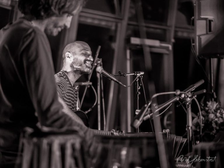 Bearded musician singing passionately at microphone with guitar during indoor concert performance – © Axel Hebenstreit