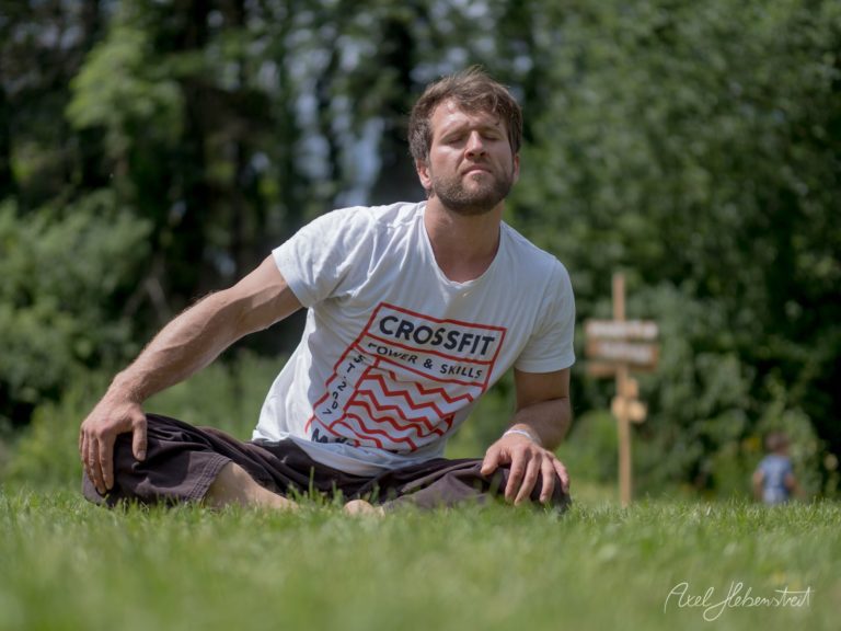 Mann in CrossFit-Shirt sitzt dehnend auf einer grünen Wiese in sonniger Natur – © Axel Hebenstreit