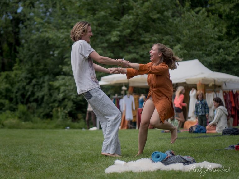 Junges Paar tanzt fröhlich auf einer Wiese bei einem Outdoor-Festival mit Zelten und Zuschauern im Hintergrund – © Axel Hebenstreit