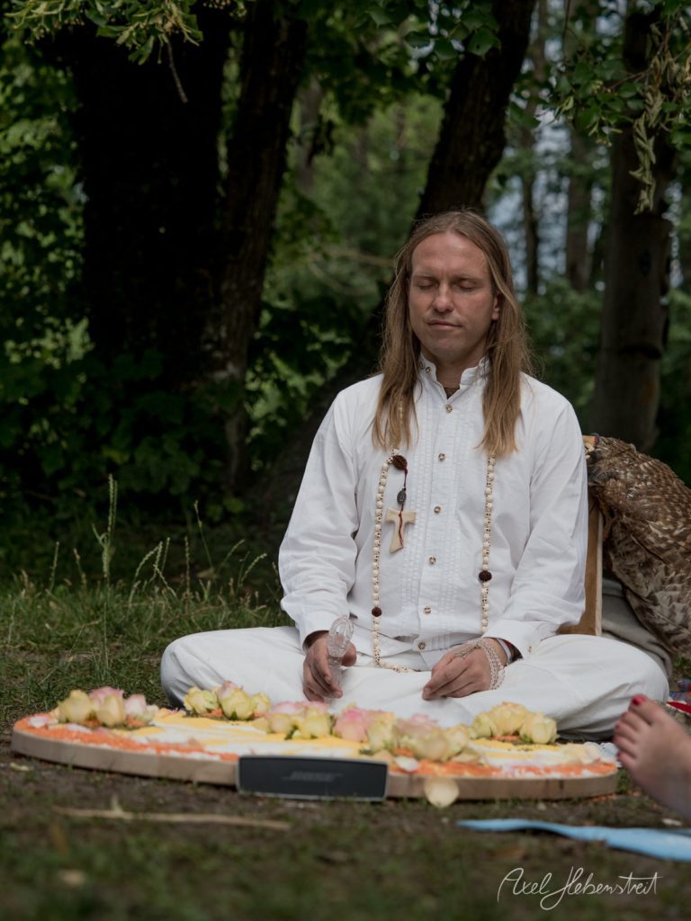 Frau in weißem Gewand meditiert friedlich im Wald neben einem bunten Blüten-Mandala – © Axel Hebenstreit