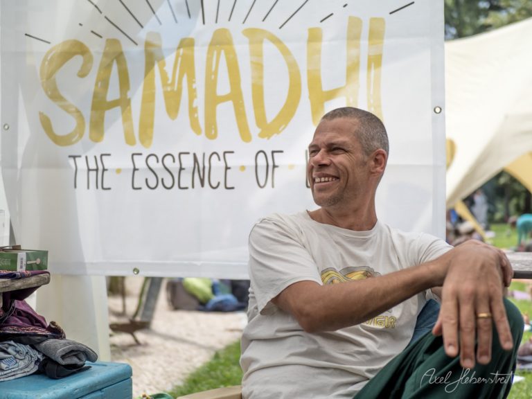 Mann in grauem T-Shirt sitzt lächelnd vor einem Schirm mit goldenem Samadhi-Schriftzug auf einem Yoga-Festival – © Axel Hebenstreit