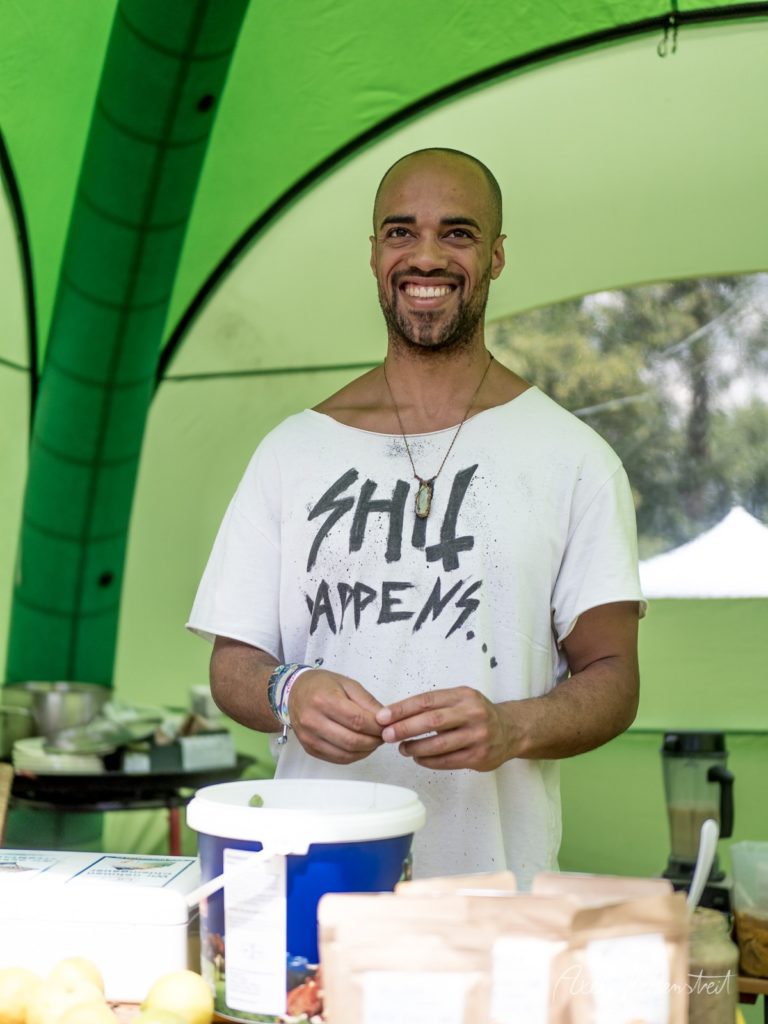 Lächelnder Mann in weißem T-Shirt bereitet Essen in grünem Festivalzelt zu – © Axel Hebenstreit