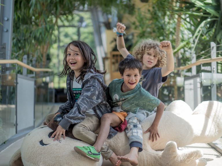 Drei fröhliche Kinder posieren jubelnd auf einer weißen Skulptur in einem Glashaus mit üppiger Bepflanzung – © Axel Hebenstreit
