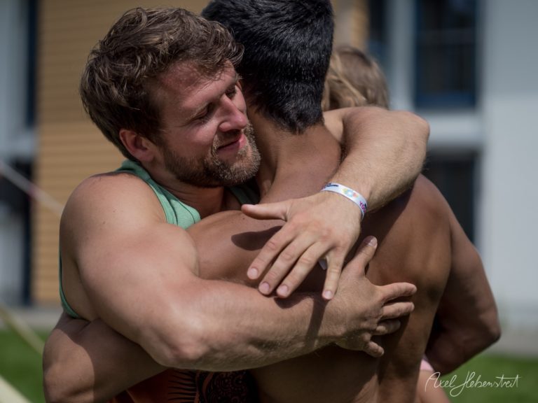 Zwei Männer umarmen sich freudig bei einem Outdoor-Event vor einem Holzgebäude – © Axel Hebenstreit