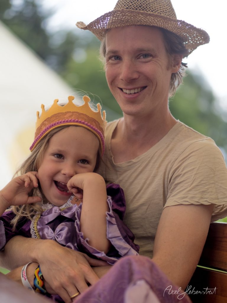 Fröhlicher Vater mit Strohhut und lachende Tochter in Prinzessinnenkostüm beim gemeinsamen Porträt im Freien – © Axel Hebenstreit