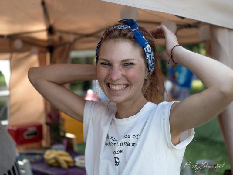 Strahlende junge Frau mit blauem Kopftuch unter einem Zeltdach auf einem Festival – © Axel Hebenstreit