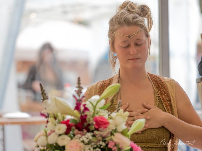 Frau in goldenem Gewand mit geschlossenen Augen hält Blumenstrauß mit rosen und lilien in hellem Raum – © Axel Hebenstreit
