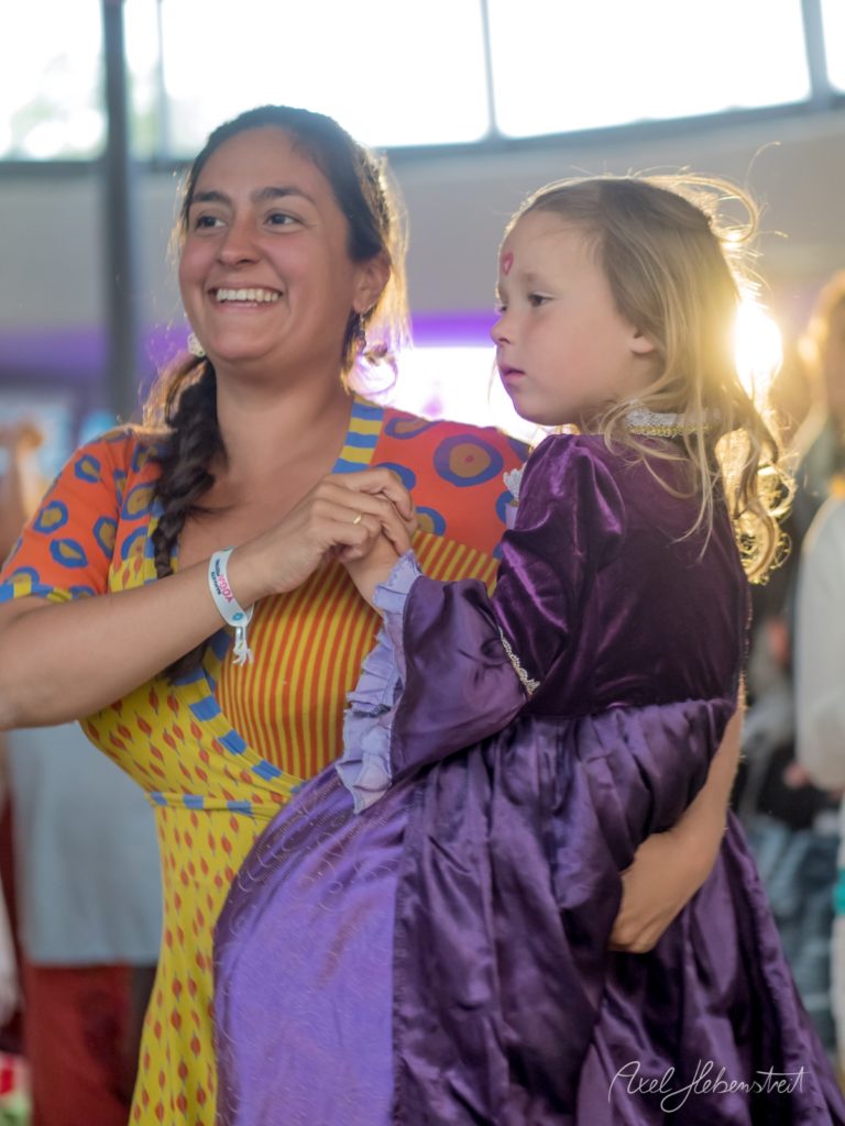 Lachende Frau in buntem Outfit hält kleines Mädchen in lila Kleid auf einem Festival – © Axel Hebenstreit