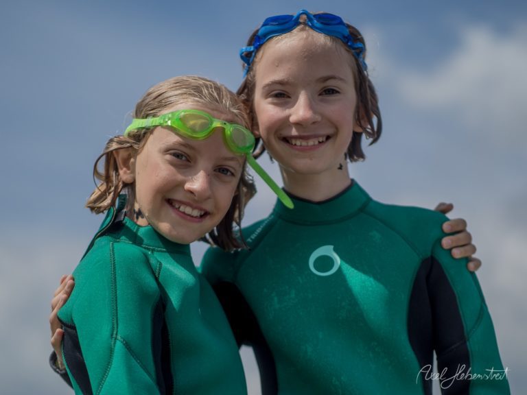 Zwei glückliche Kinder in grünen Neoprenanzügen mit Schwimmbrillen strahlen vor blauem Himmel – © Axel Hebenstreit