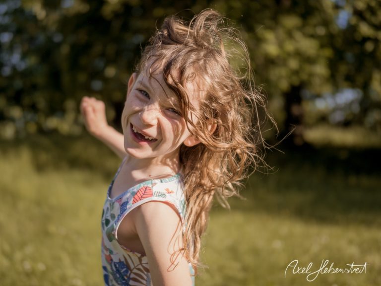 Ein fröhliches Kind mit wildem Haar dreht sich lachend im Grünen, umgeben von natürlicher Schönheit. – © Axel Hebenstreit