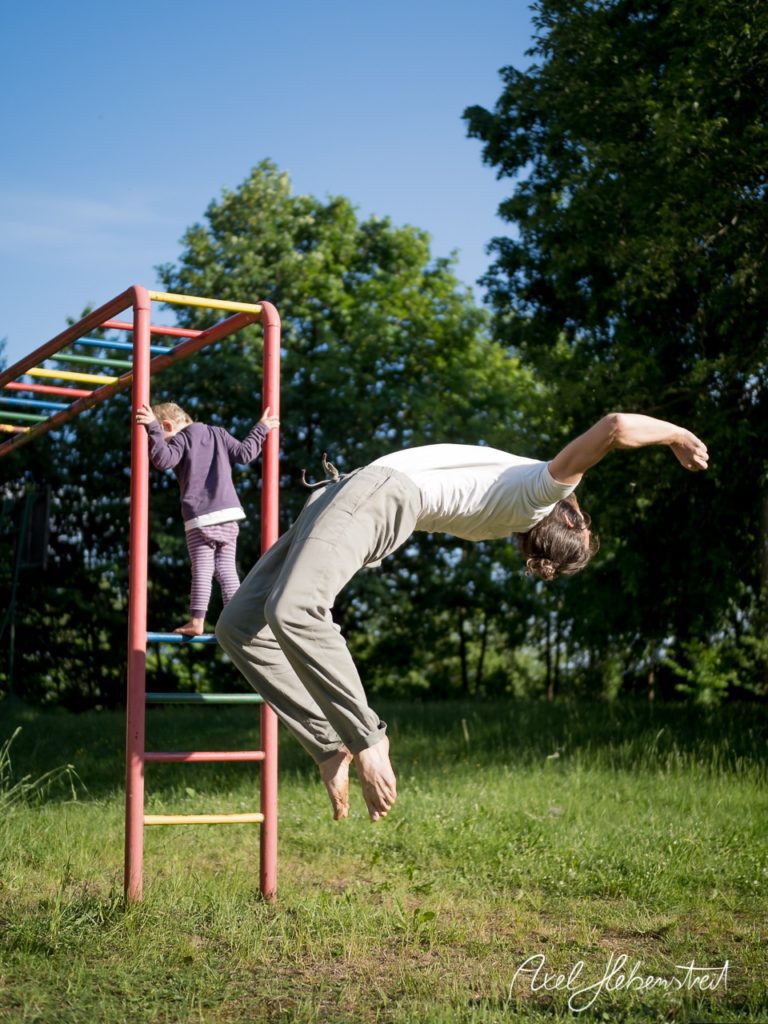 Ein Mann springt fröhlich von einer Stange, während ein Kind im Hintergrund daran klettert, umgeben von Natur. – © Axel Hebenstreit