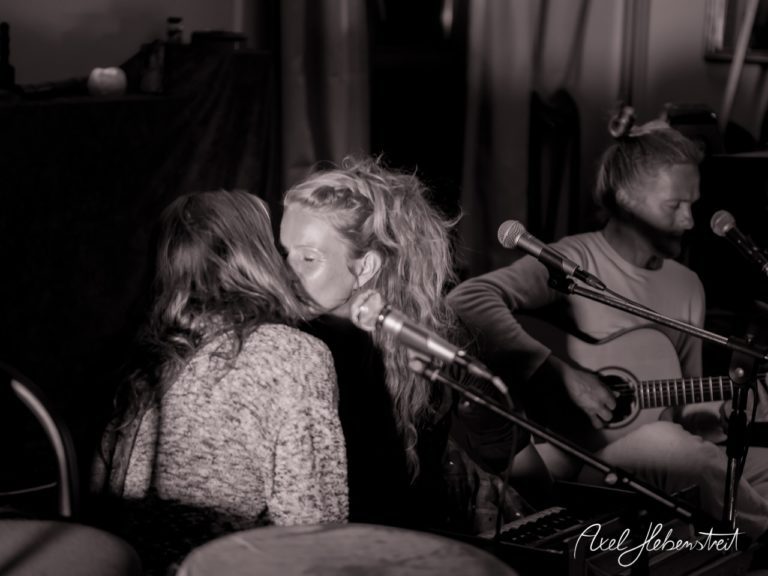 Zwei Frauen küssen sich intim während eines akustischen Konzerts, während ein Musiker im Hintergrund spielt. – © Axel Hebenstreit