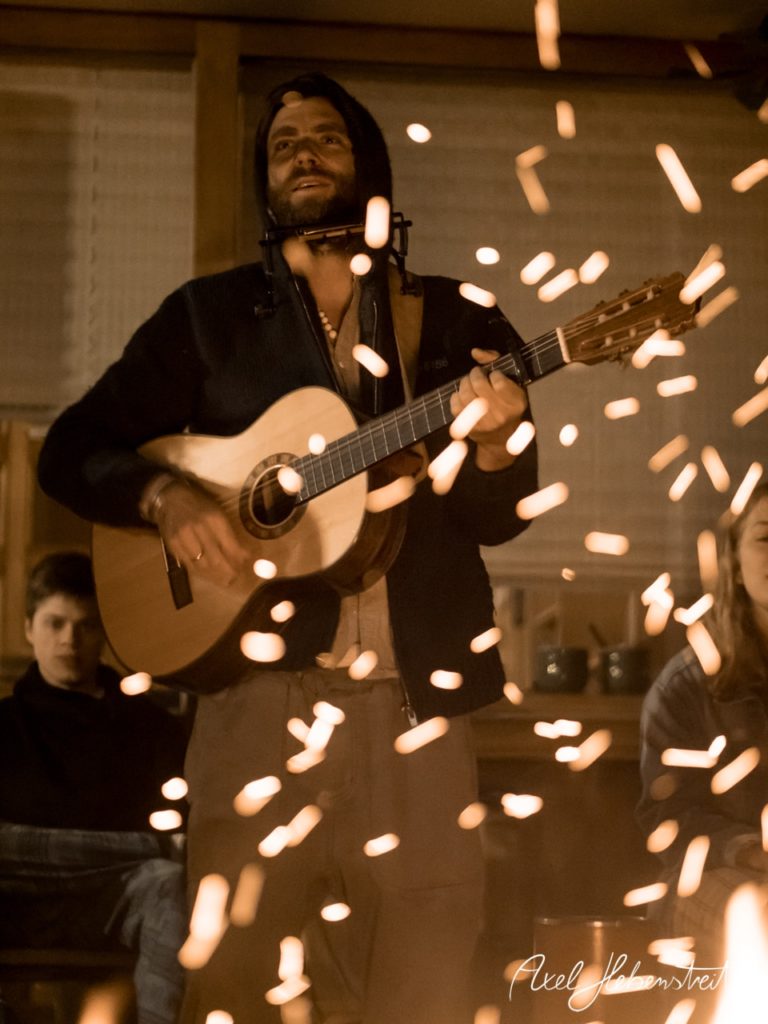 Ein Musiker spielt Gitarre vor einem Lagerfeuer, während Zuschauer gebannt lauschen. – © Axel Hebenstreit