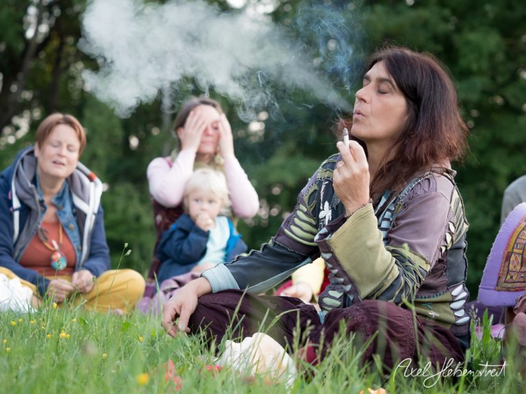 Eine Gruppe von Menschen meditiert im Freien, während eine Frau Rauch aus einem Räucherstäbchen ausstößt. – © Axel Hebenstreit