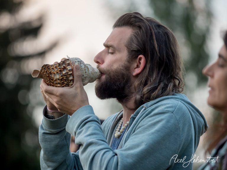 Ein Mann mit Bart bläst in eine Muschel und erzeugt eine harmonische Atmosphäre im Freien. – © Axel Hebenstreit