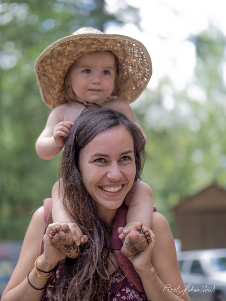 Mutter trägt lachend ihre kleine Tochter mit Strohhut auf den Schultern in grüner Gartenumgebung – © Axel Hebenstreit