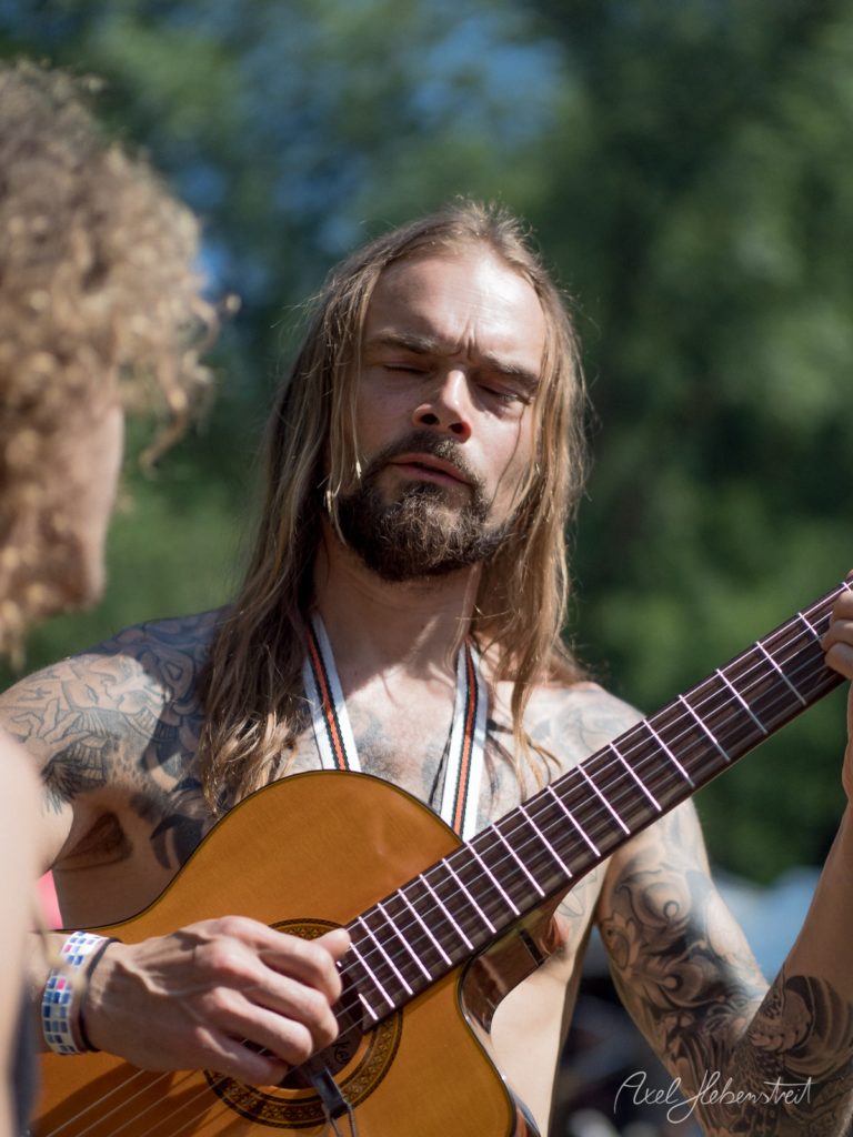 Tätowierter Musiker mit langen Haaren spielt konzentriert Akustikgitarre im Freien bei Sonnenschein – © Axel Hebenstreit
