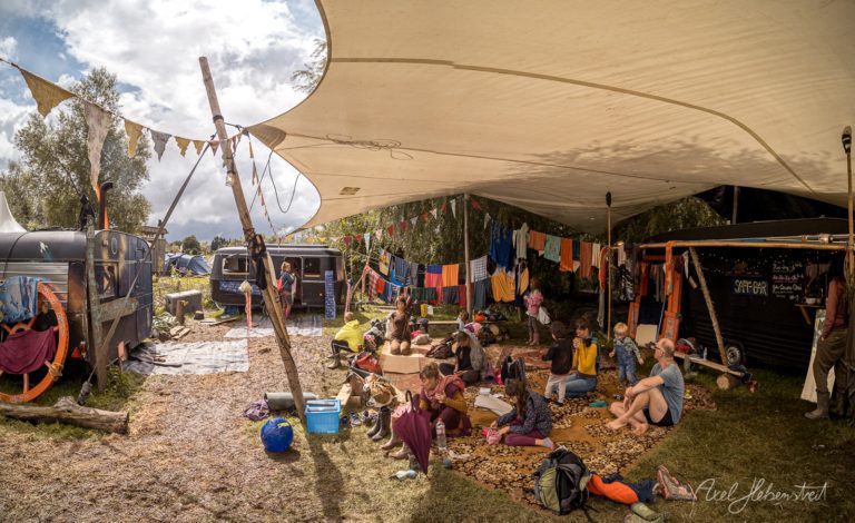 Festivalcommunity sitzt unter Sonnensegel neben Wohnwagen und Zelten bei gemeinsamem Lagerfeuer-Treffen – © Axel Hebenstreit