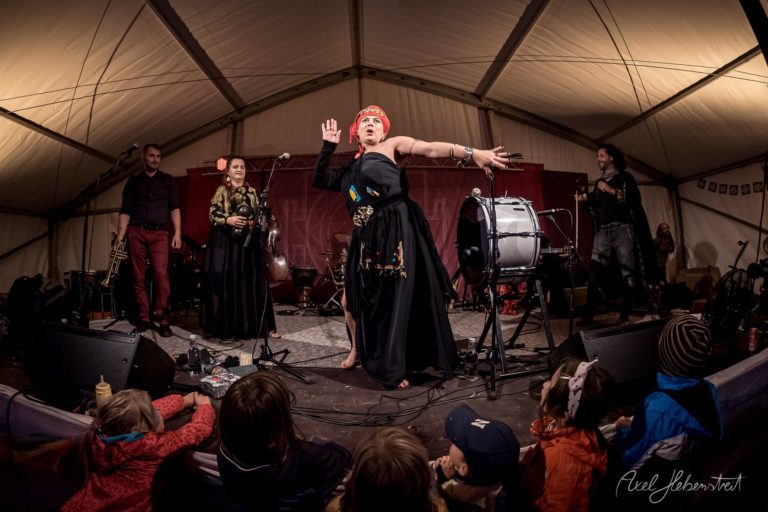 Sängerin in roter Kappe und schwarzem Gewand performt ausdrucksstark unter Zeltdach vor sitzendem Publikum – © Axel Hebenstreit