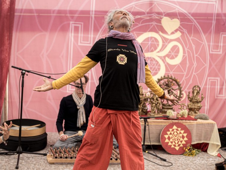 Ein Mann mit weißem Haar und Bart performt dynamisch auf einer Yoga-Bühne mit pinkem Hintergrund und spirituellen Symbolen – © Axel Hebenstreit
