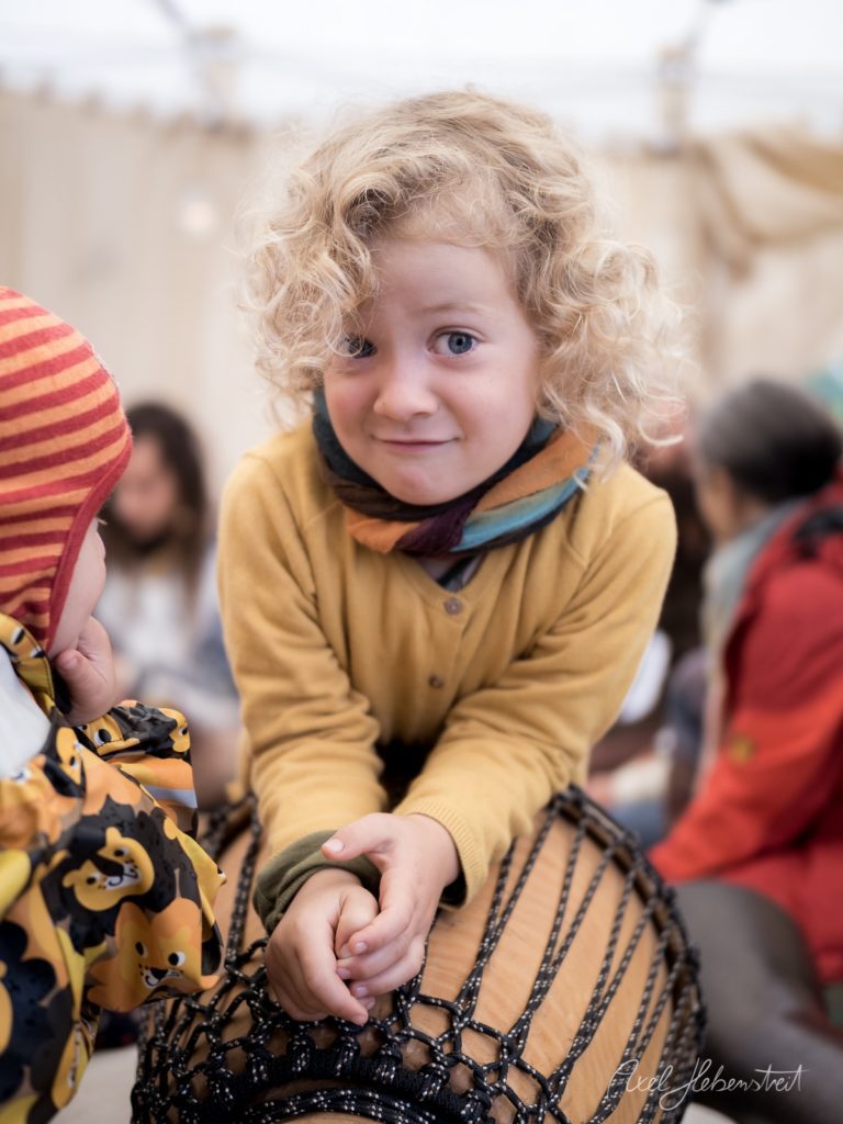 Lockiges Kleinkind in gelbem Pullover hält lächelnd ein buntes Drachenseil auf einem Fest – © Axel Hebenstreit