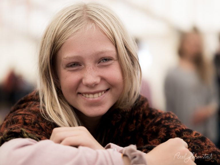 Lächelndes Mädchen mit blondem Haar in braunem Pullover vor unscharfem Hintergrund – © Axel Hebenstreit