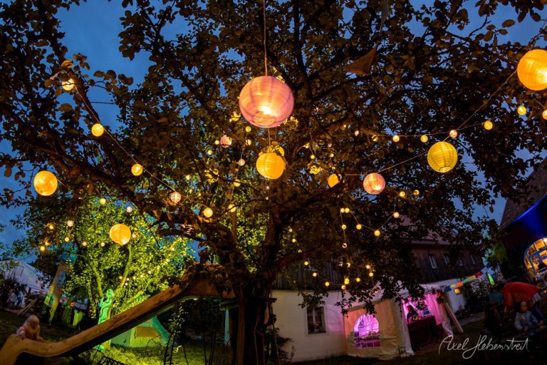 Festlich illuminierter alter Baum mit gelben und rosa Lampions bei Nacht auf einem Festivalgelände in Bayern – © Axel Hebenstreit