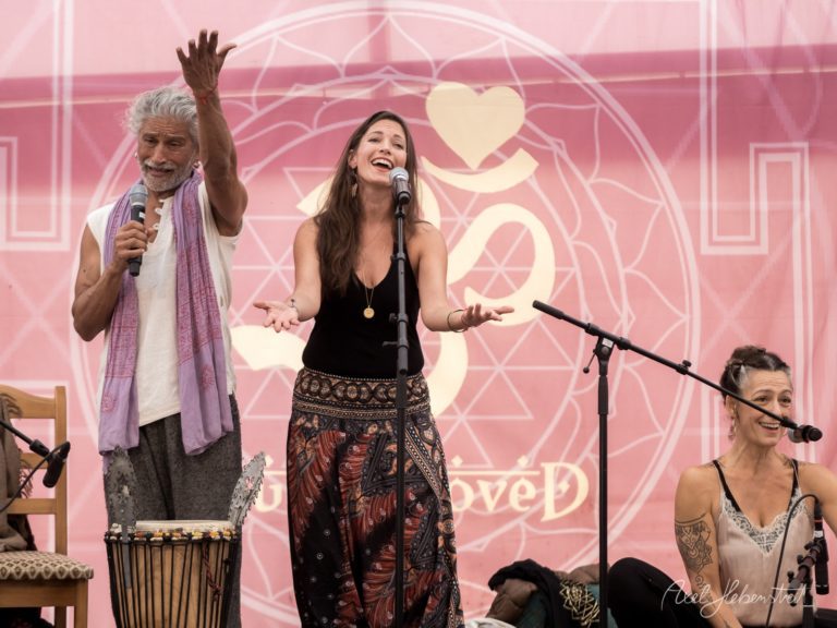 Drei Musiker treten auf einer Bühne mit rosa Hintergrund und Herzdesign auf, einer spielt Djembe, während die beiden Frauen singen – © Axel Hebenstreit