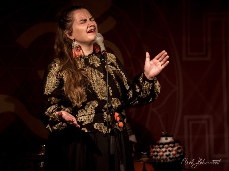 Sängerin in goldenem Brokatgewand performt emotional am Mikrofon auf dunkler Bühne – © Axel Hebenstreit