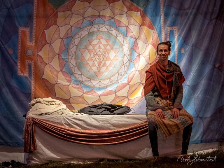 Junge Frau sitzt lächelnd vor einem farbenfrohen Mandala-Wandteppich auf einem drapierten Tisch bei einem Festival – © Axel Hebenstreit